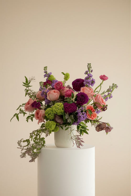 IN𝓃 Floral Studio Blush Atelier arrangement with pink roses, purple dahlias, green hydrangeas, and purple stock flowers in a white vase.