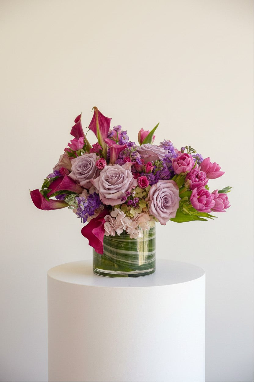 IN𝓃 Floral Studio Blush Calla arrangement with pink calla lilies, lavender roses, pink tulips, and hydrangeas in a glass vase.