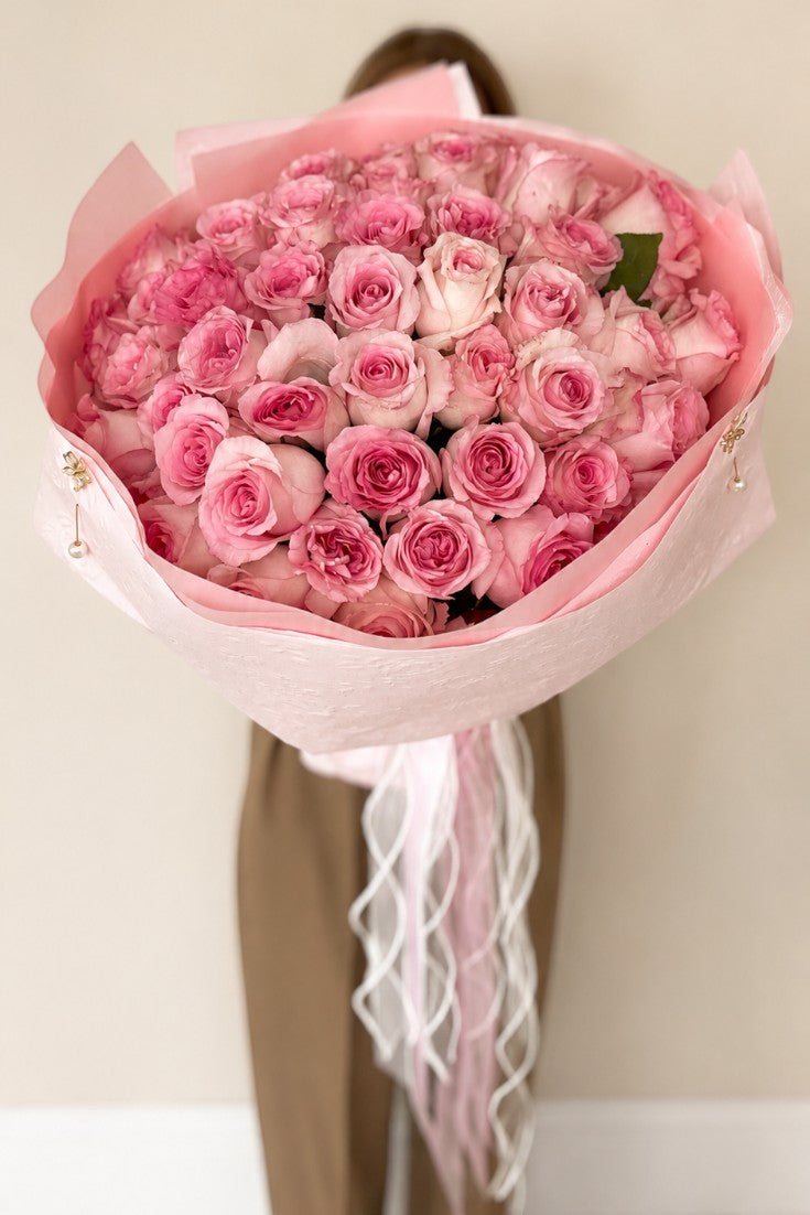 IN𝓃 Floral Studio blush pink rose bouquet wrapped in pink paper with pearl accents and flowing ribbons held by a person