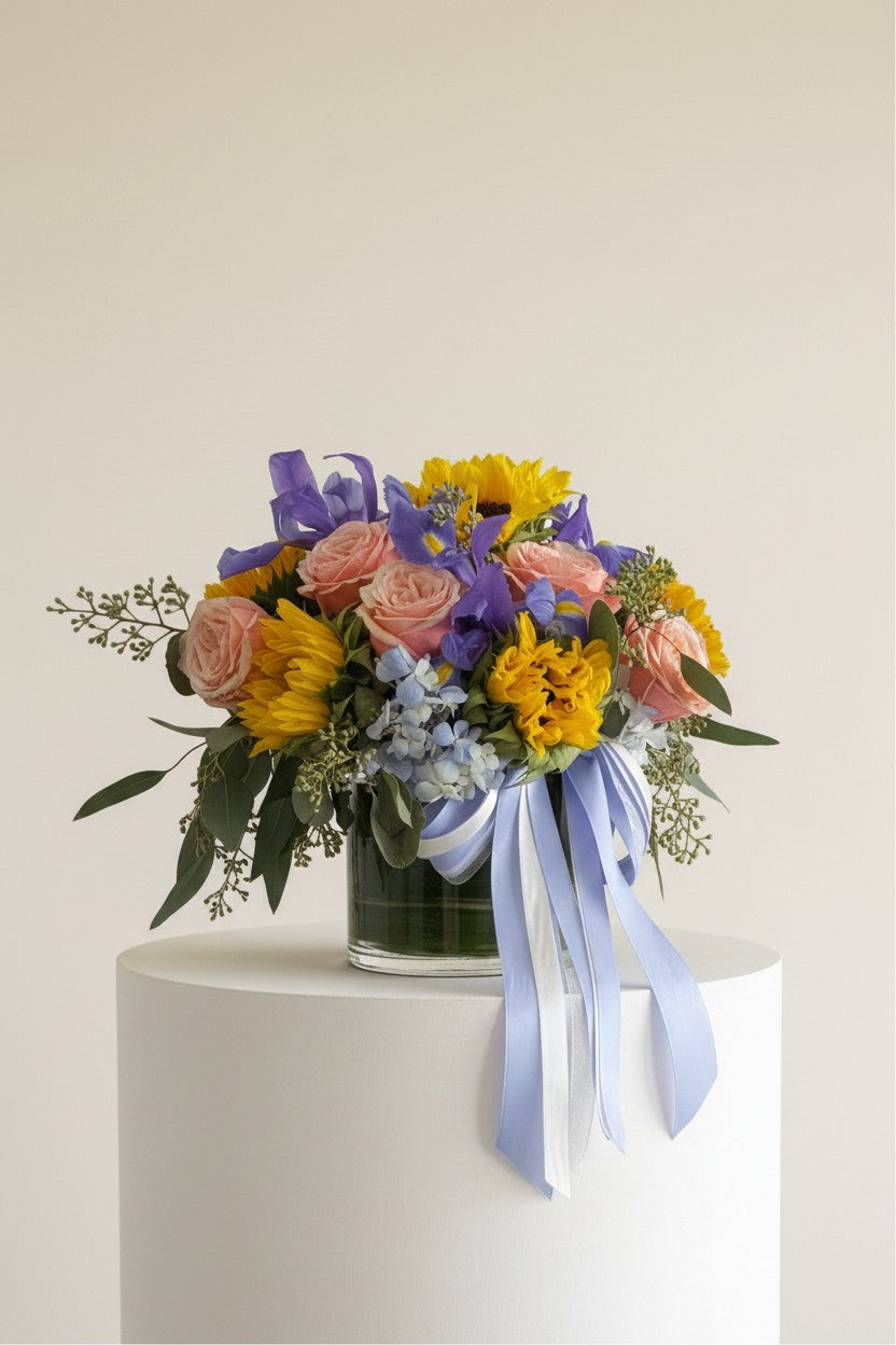 IN𝓃 Floral Studio Golden Garden arrangement with pink roses, yellow sunflowers, purple irises, and blue hydrangeas in a glass vase with light blue and silver ribbons.