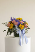 IN𝓃 Floral Studio Golden Garden arrangement with pink roses, yellow sunflowers, purple irises, and blue hydrangeas in a glass vase with light blue and silver ribbons.