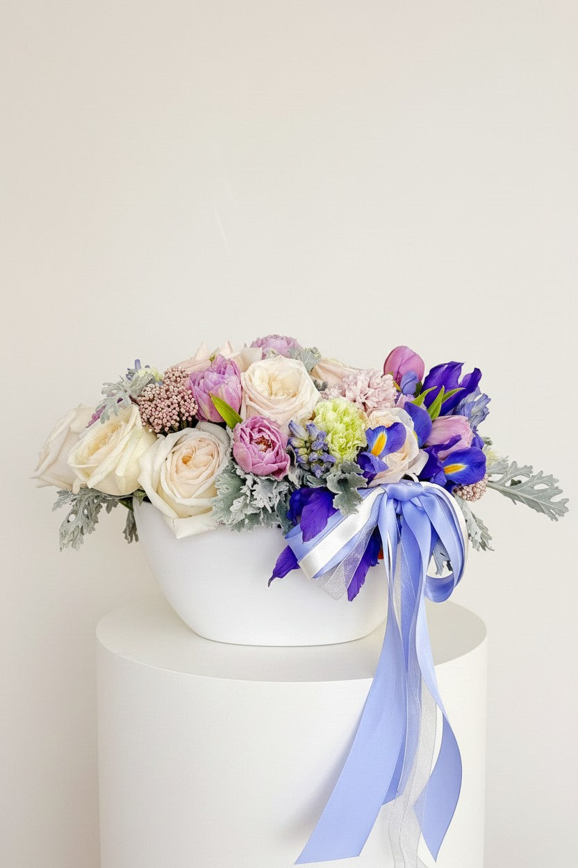 IN𝓃 Floral Studio Lavender Whisper arrangement: white roses, purple tulips, blue irises, and greenery in a white ceramic bowl with blue ribbons.