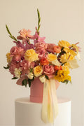 IN𝓃 Floral Studio pastel garden arrangement with blush pink roses, yellow roses, ranunculus, and gladiolus in a pink hat box with ribbons.