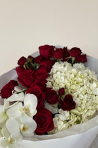 IN𝓃 Floral Studio red roses, white orchids, and hydrangeas bouquet wrapped in white paper with a white ribbon.