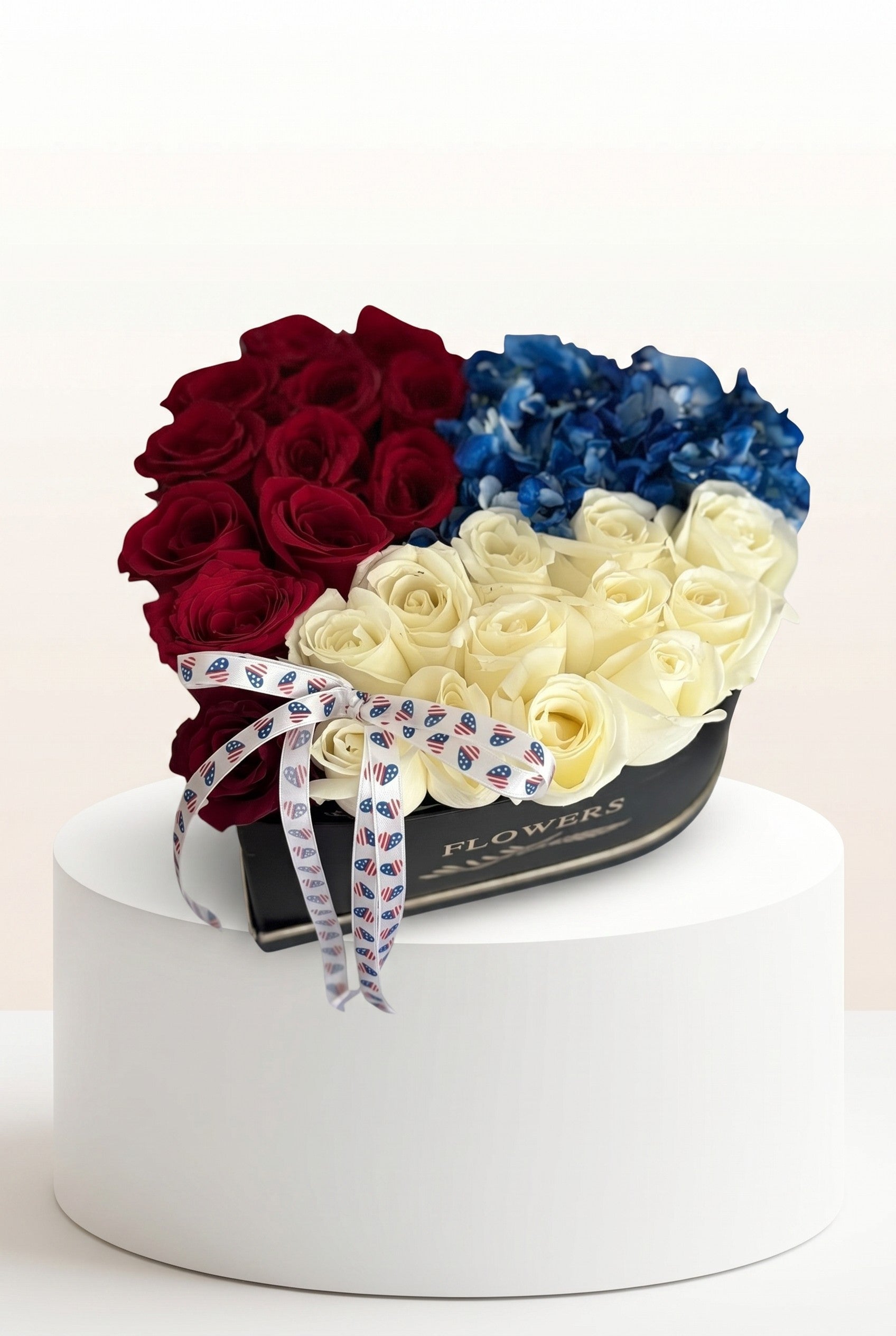 IN𝓃 Floral Studio heart-shaped box with red roses, white roses, and blue hydrangeas, finished with a patriotic ribbon.