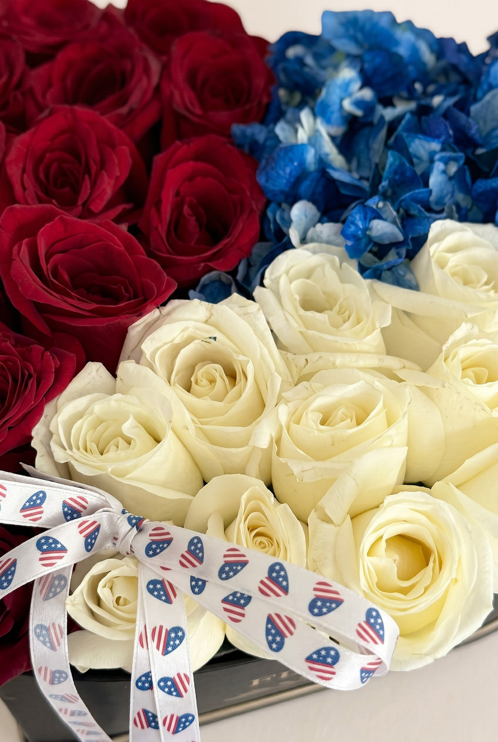 IN𝓃 Floral Studio patriotic heart-shaped box with red and white roses, blue hydrangeas, and a heart-patterned ribbon.